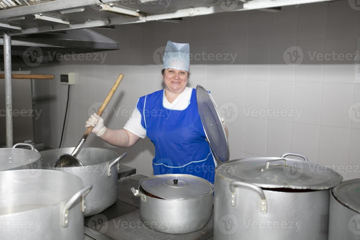 en kvinna laga mat i ett industriell kök med en stor slev bland metall kokkärl. kock på de sjukhus foto
