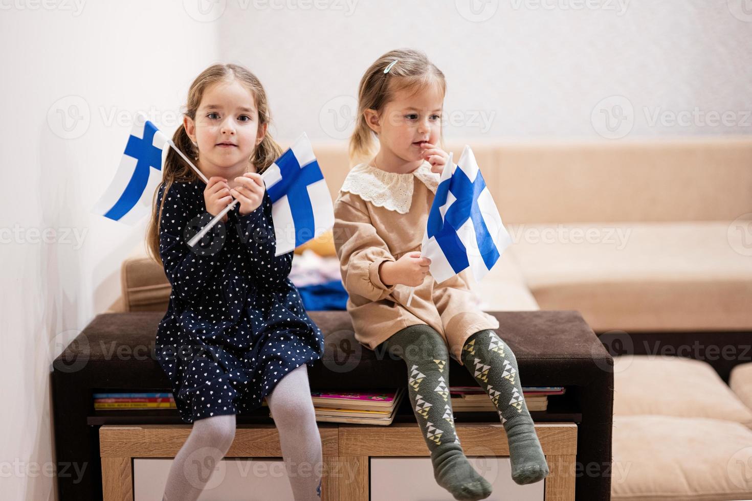 två systrar är Sammanträde på en soffa på Hem med finska flaggor på händer. finland barn flickor med flagga . foto