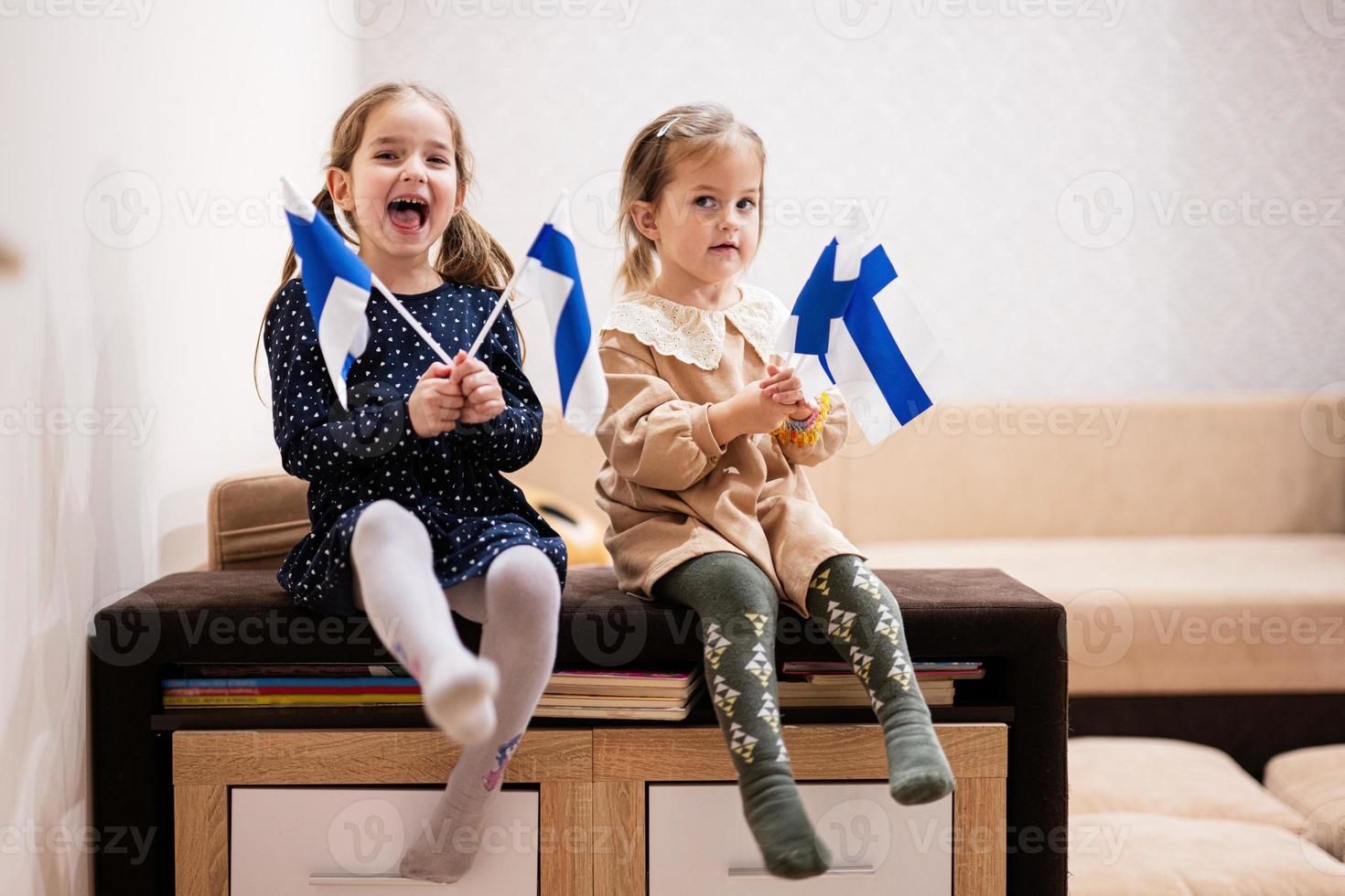 två systrar är Sammanträde på en soffa på Hem med finska flaggor på händer. finland barn flickor med flagga . foto