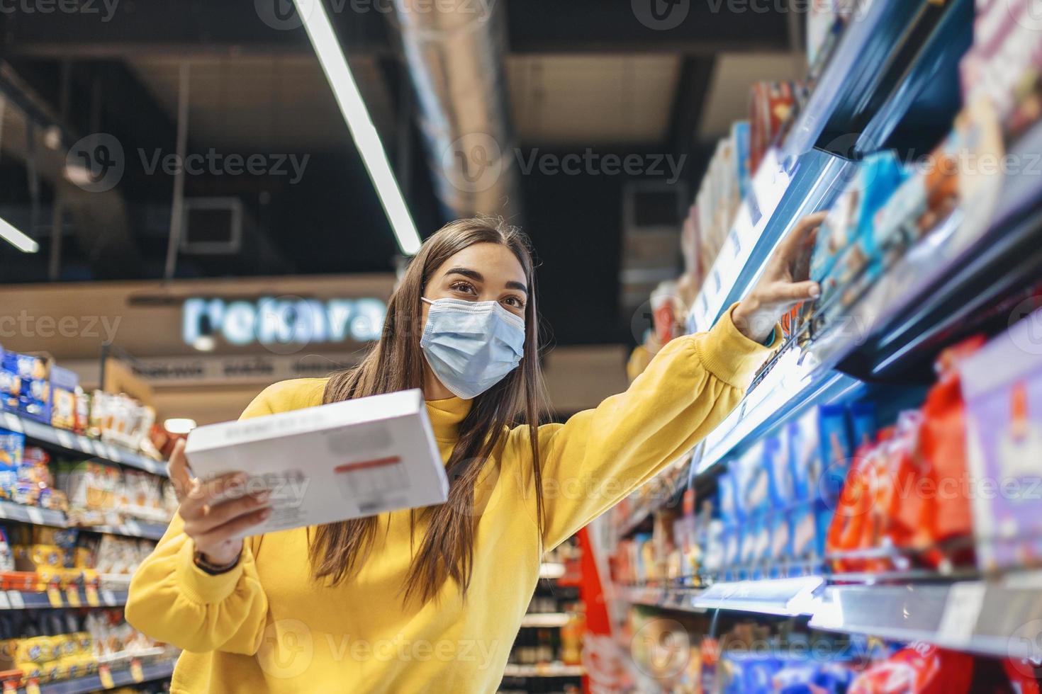 social avstånds i en mataffär. en ung kvinna i en disponibel ansikte mask uppköp mat och sätta dem i en matvaror korg. handla under de coronavirus covid-19 epidemi foto