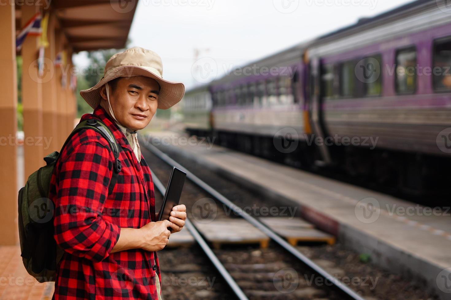 asiatisk man resande är på järnväg station, bär hatt, innehar smart läsplatta. begrepp, resa förbi tåg i thailand kan bok eller förbi biljett uppkopplad. teknologi och transport. foto