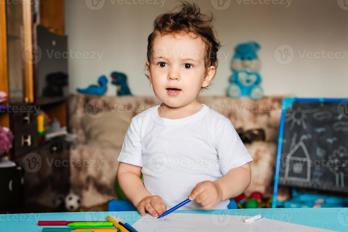 en små pojke drar på ark av papper liggande på de tabell med färgad pennor foto