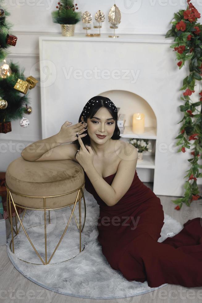 porträtt av Söt ung flicka mysigt sitta ner på de golv, leende ha på sig röd klänning i dekorerad jul levande rum inomhus foto