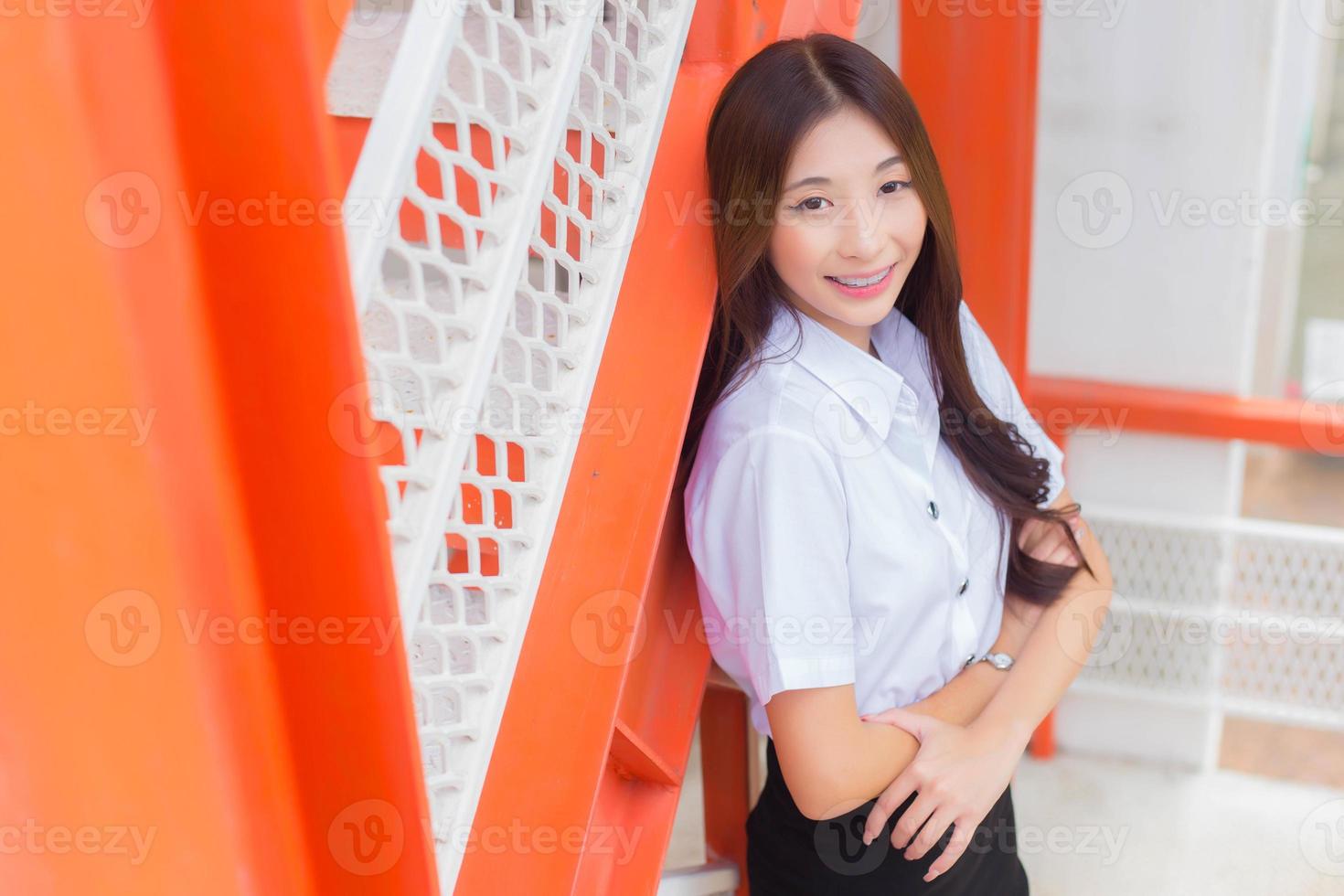 porträtt av ett vuxen thai studerande i universitet studerande enhetlig. asiatisk skön flicka stående leende lyckligt var henne tänder är bär hållare ser på kamera självsäkert till närvarande något. foto