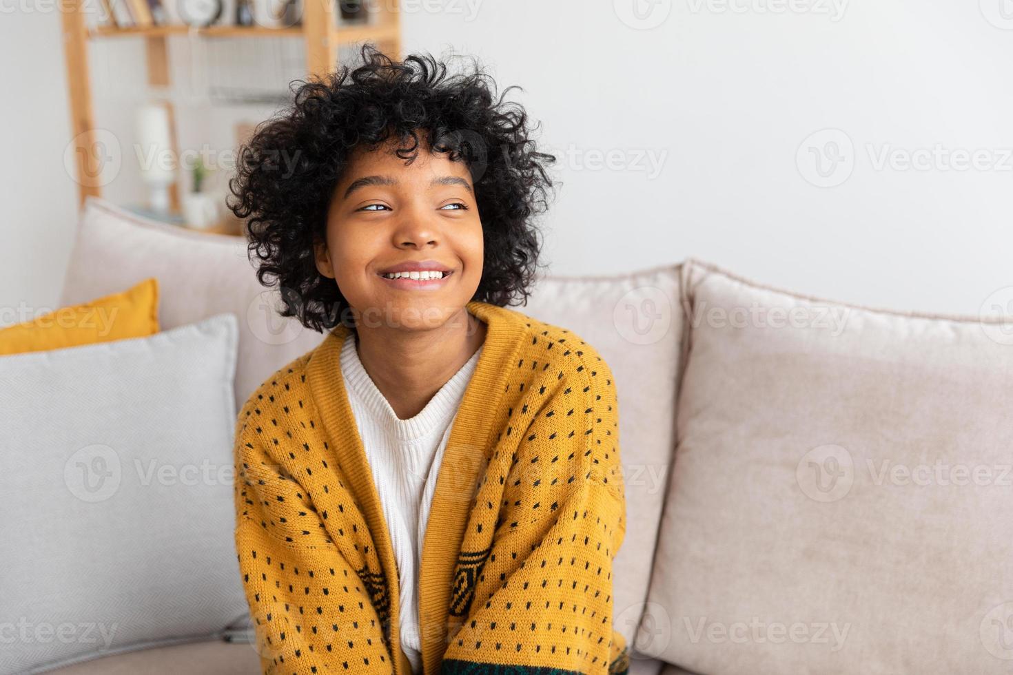 skön afrikansk amerikan flicka med afro frisyr leende Sammanträde på soffa på Hem inomhus. ung afrikansk kvinna med lockigt hår skrattande. frihet lycka sorglös Lycklig människor begrepp. foto