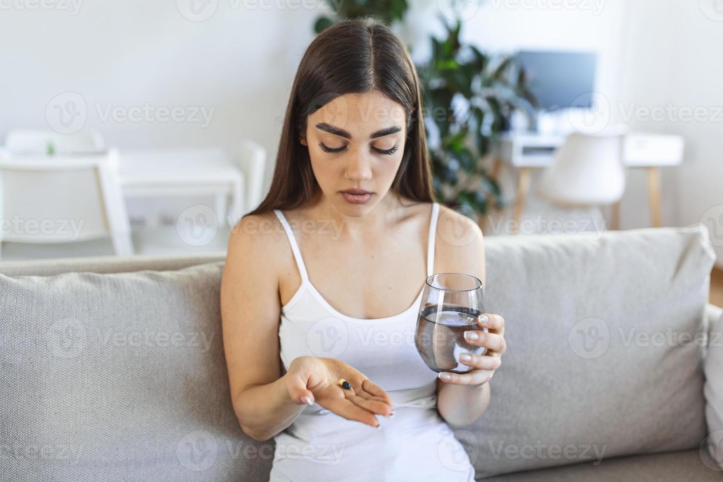 ung kvinna tar piller med glas av vatten i hand. betonade kvinna ser i fönster och dricka nersövd antidepressivt medel medicin. kvinna känner deprimerad, tar läkemedel. mediciner på arbete foto