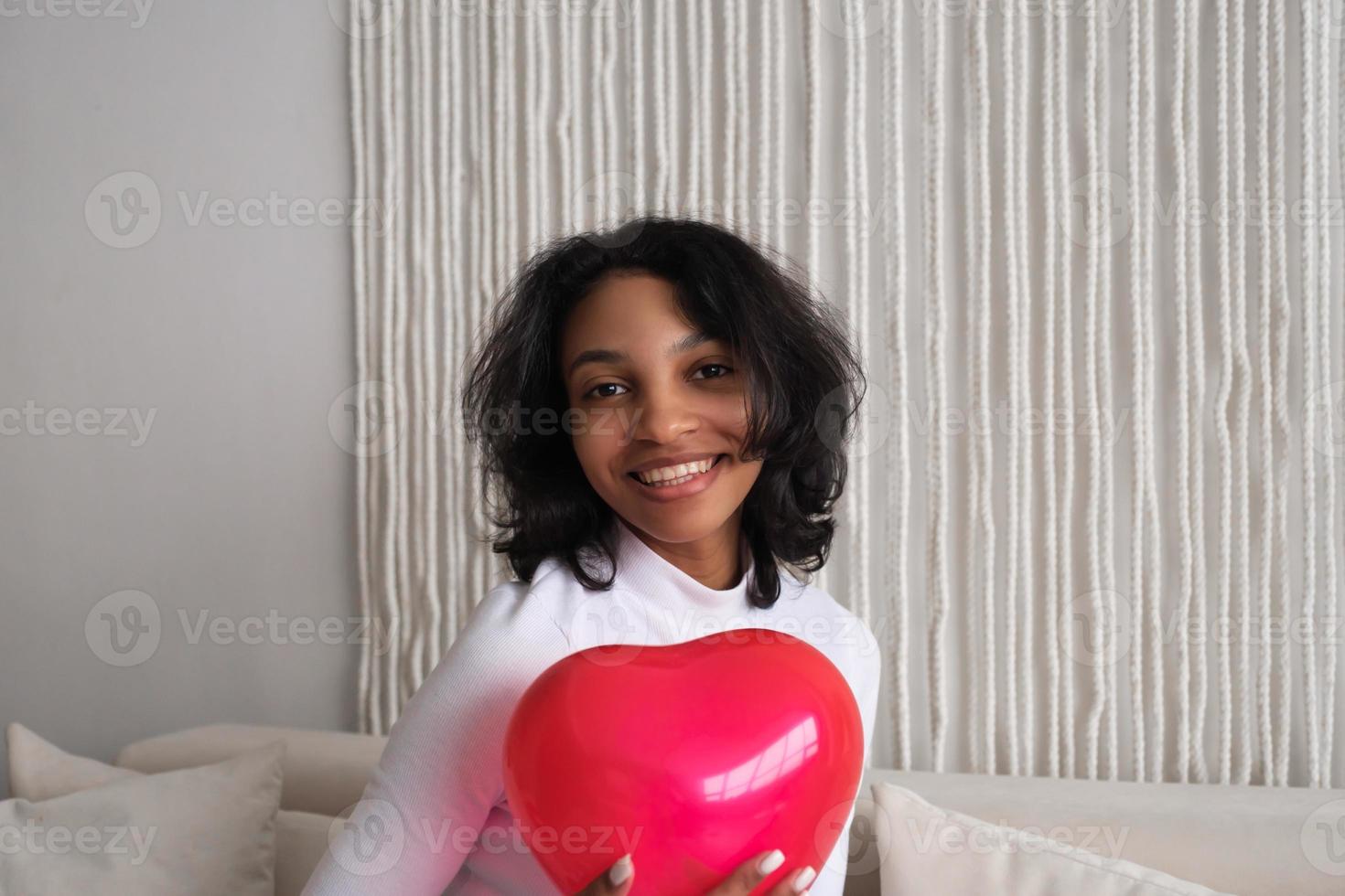 närbild porträtt av ung afrikansk amerikan kvinna innehav röd hjärta formad ballong Sammanträde på soffa på Hem. fira alla hjärtans dag, kärlek dag, kvinnors dag, födelsedag. foto