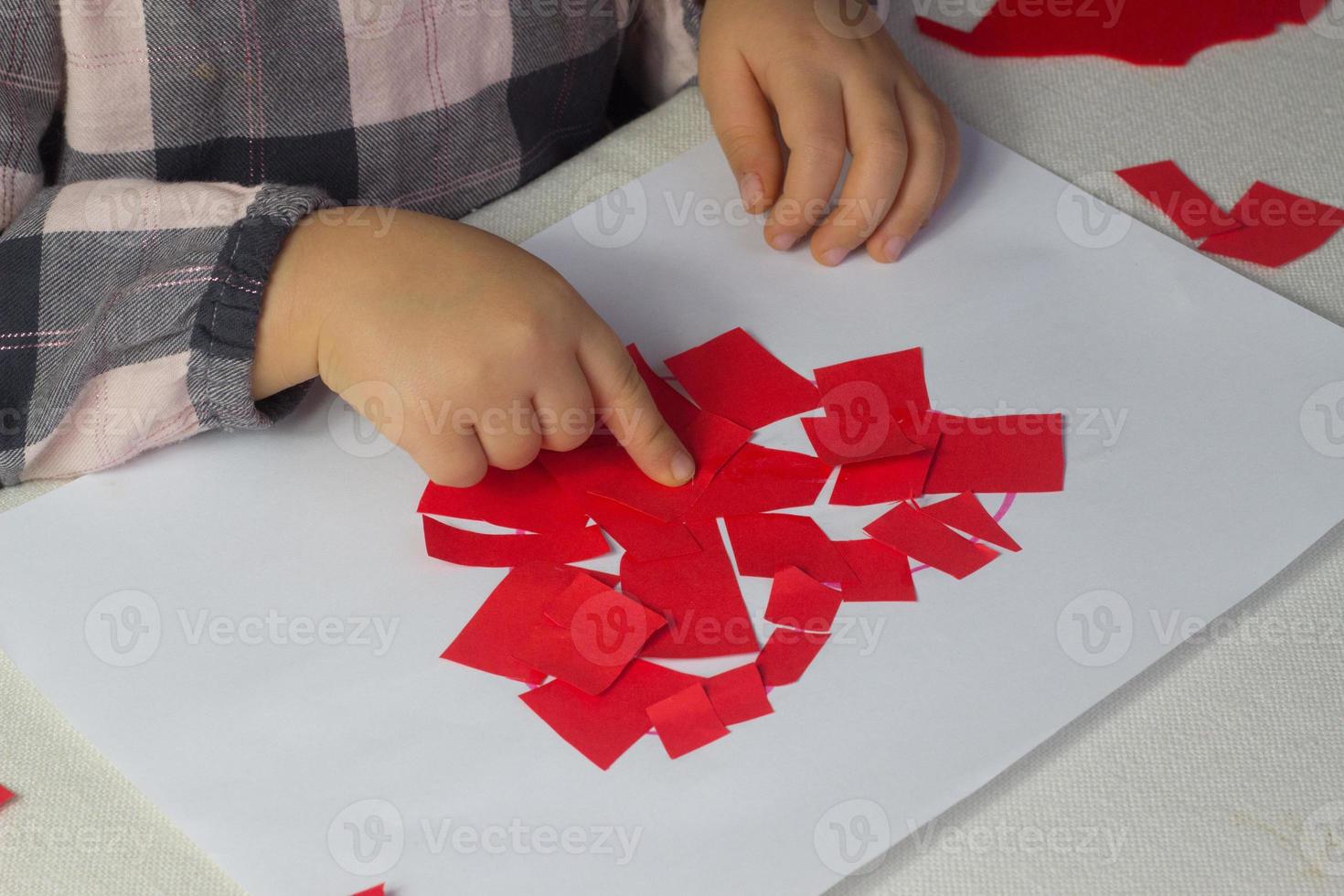 liten flicka framställning röd hjärta applique med papper och lim på vit lakan, alla hjärtans dag dag hälsning kort, Semester gåva för mödrar dag, hand tillverkad barn närvarande, kärlek symbol med papper, småbarn aktivitet foto