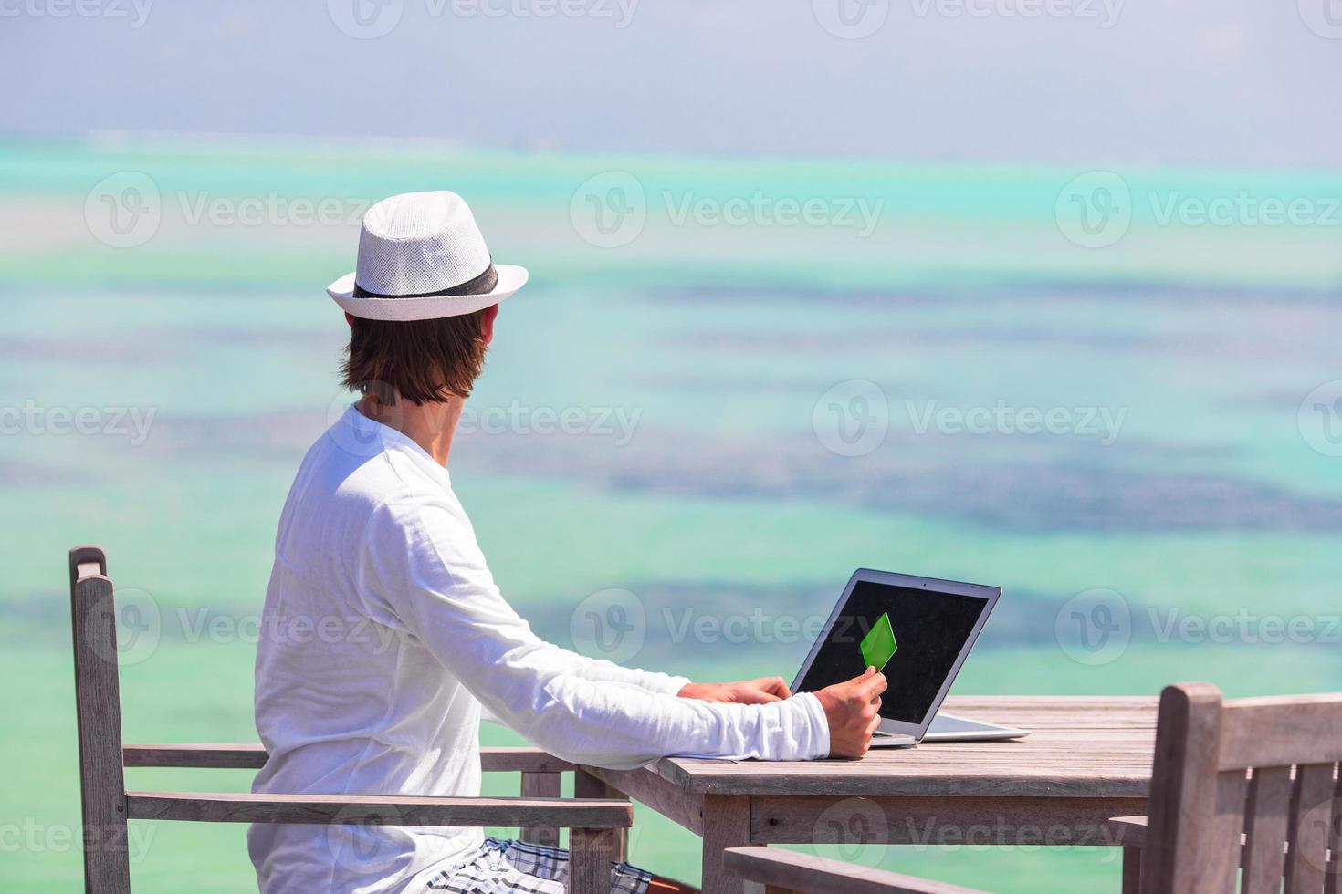 ung man arbetssätt på bärbar dator med kreditera kort på tropisk strand foto