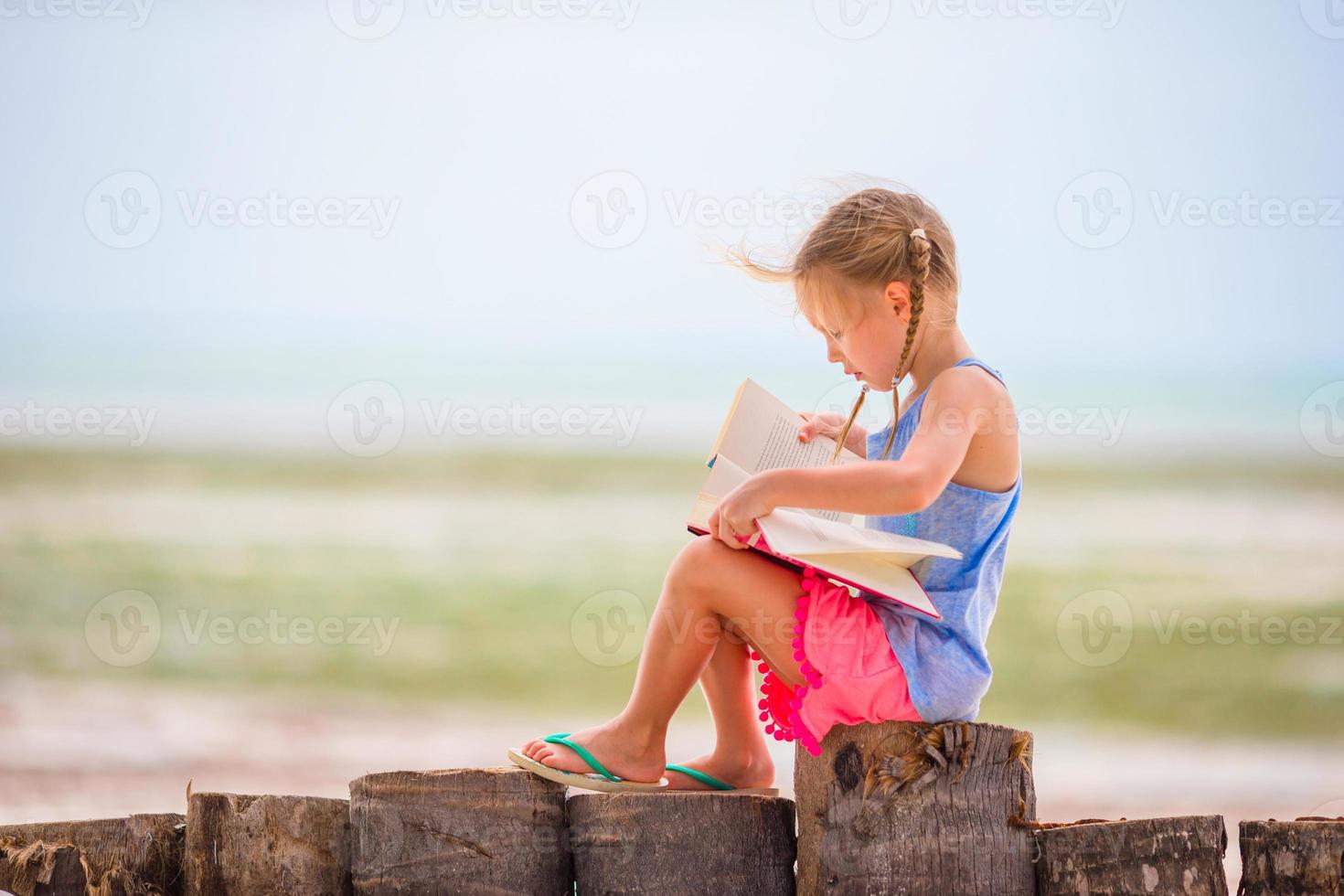 liten bedårande flicka som läser bok under den tropiska vita stranden foto