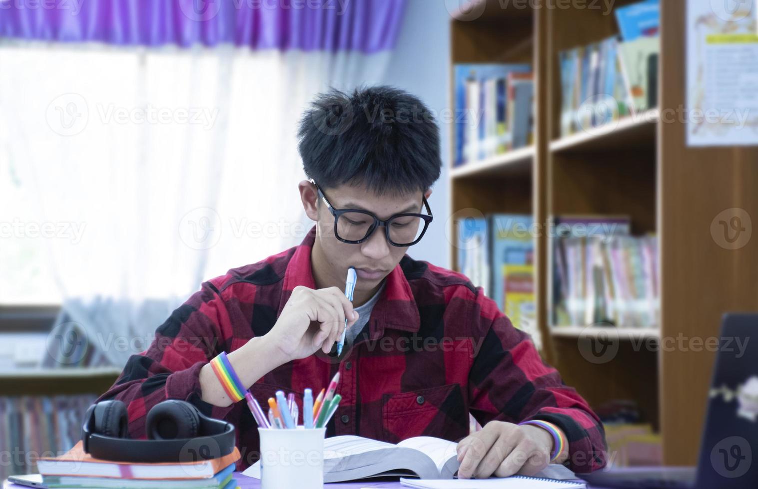 porträtt ung asiatisk pojke bär glasögon och regnbåge armband, innehav penna, Sammanträde i bibliotek, läsning böcker och koncentrering till bok på tabell innan mitten termin testa och slutlig testa Nästa dag. foto