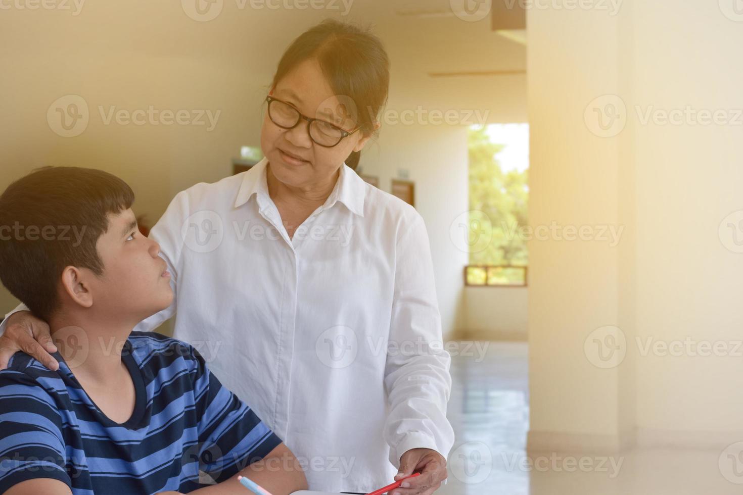 ung asiatisk pojke är håller på med hans uppdrag och lyssnande till hans kvinna äldre lärarens råd handla om skola projekt, vuxen hjälper barn till do skola projekt begrepp. foto