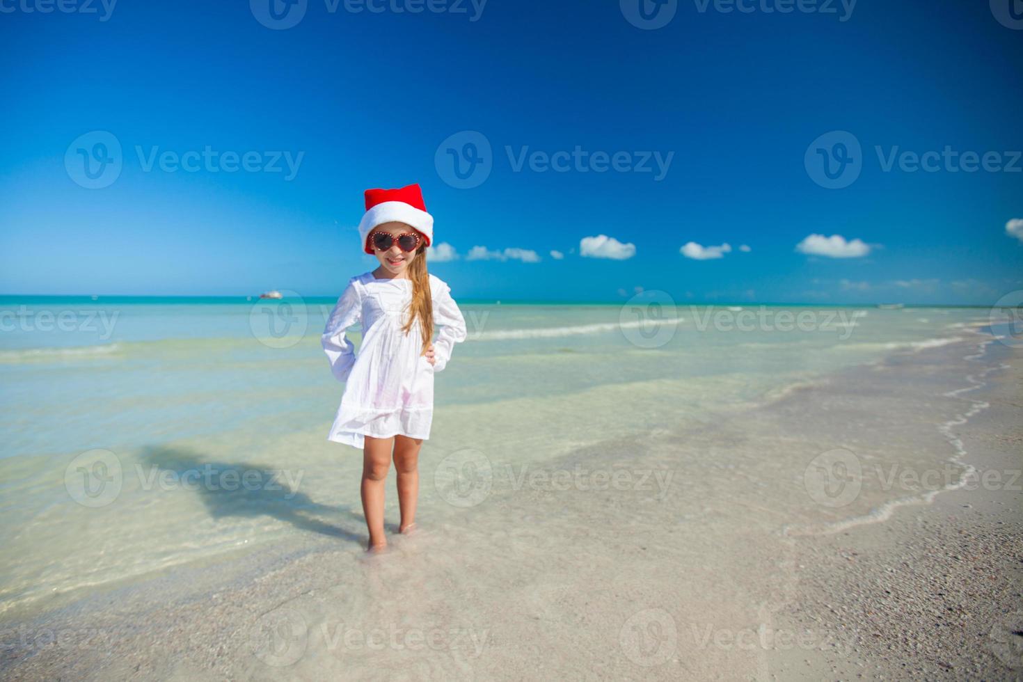 liten flicka i röd hatt santa claus och solglasögon på de strand foto