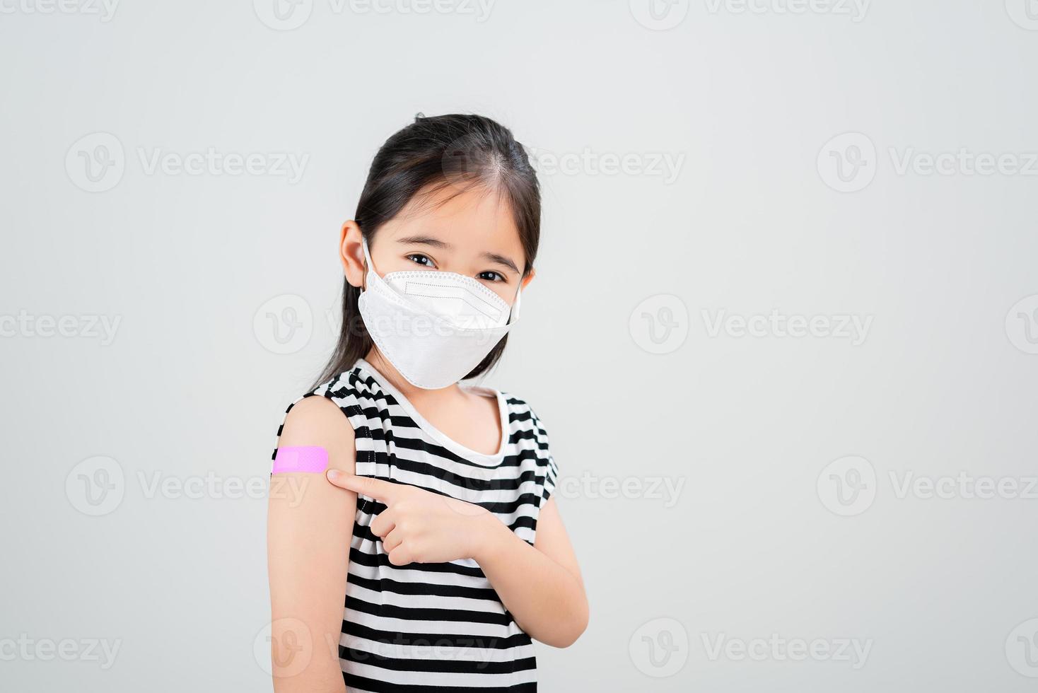 asiatisk liten flicka med medicinsk mask som visar henne ärm med bandage efter virus vaccin. unge och barn vaccination kampanj. skydda din unge med vaccin begrepp foto