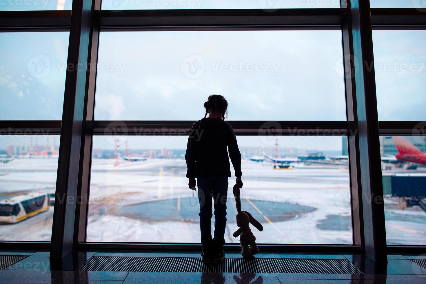 liten flicka i flygplats nära stor fönster medan vänta för ombordstigning foto