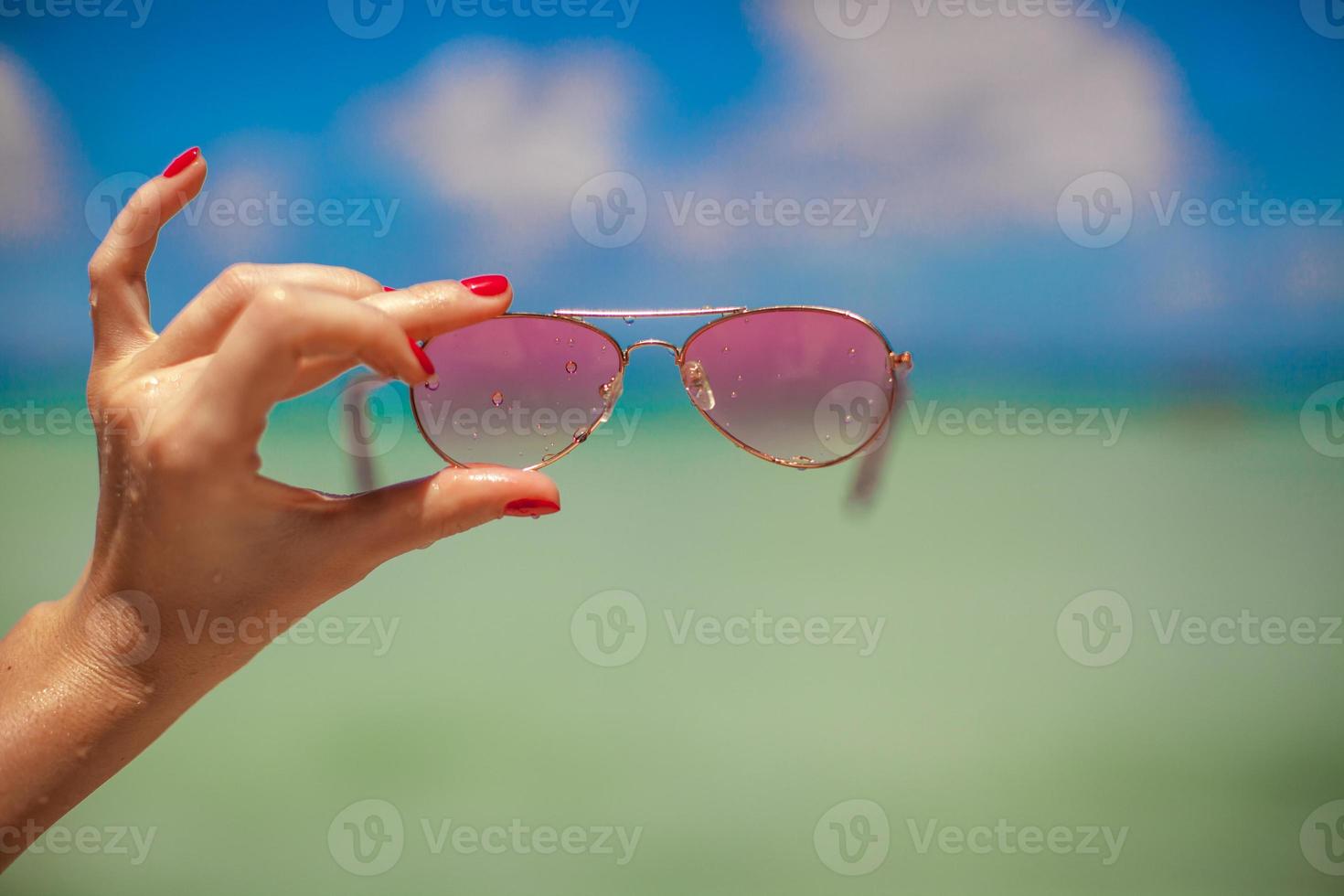 kvinnas hand innehav rosa solglasögon på tropisk strand bakgrund de hav foto
