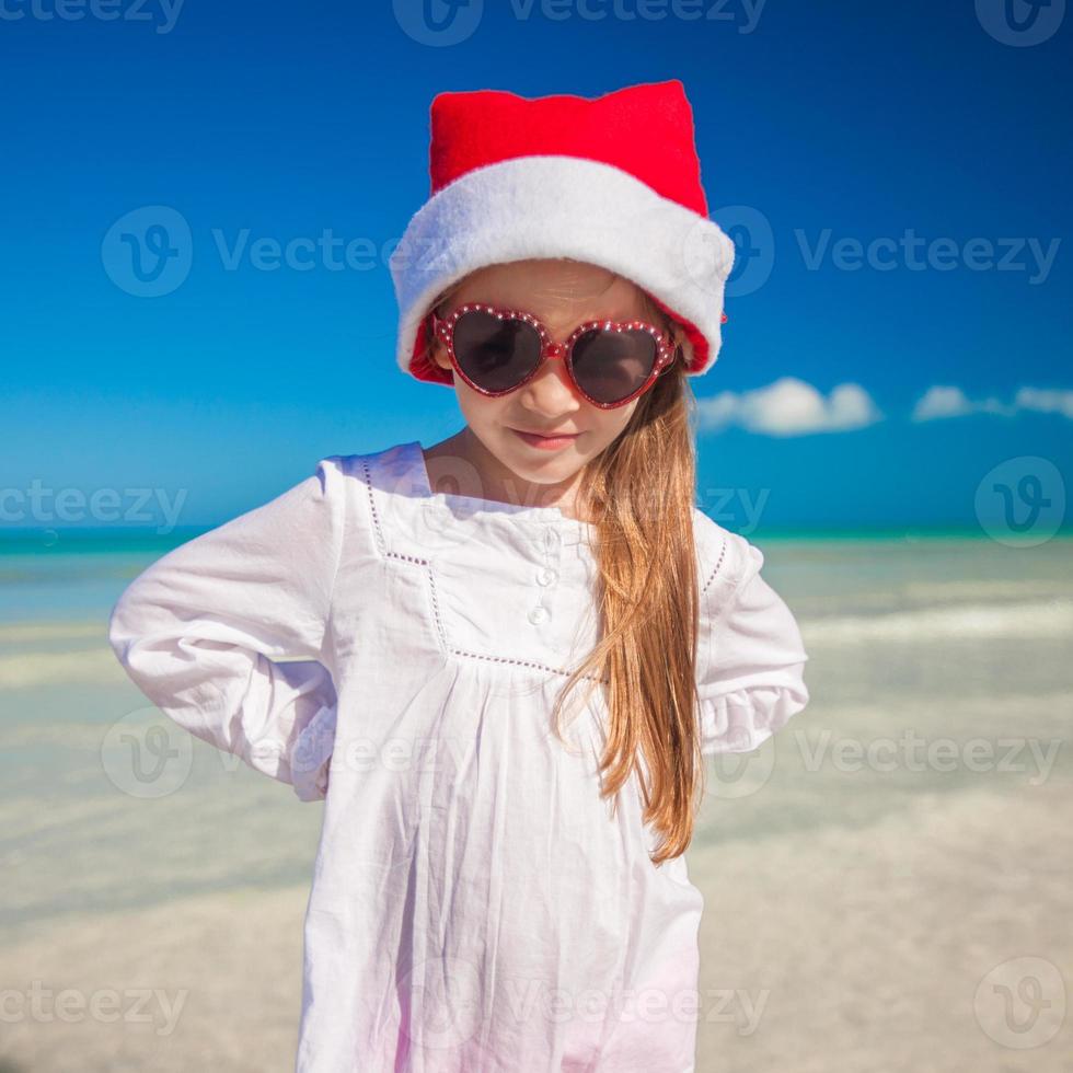 liten förtjusande flicka i röd santa hatt på tropisk strand foto