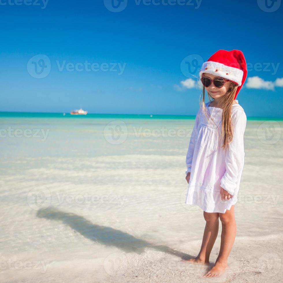 liten förtjusande flicka i röd santa hatt på tropisk strand foto