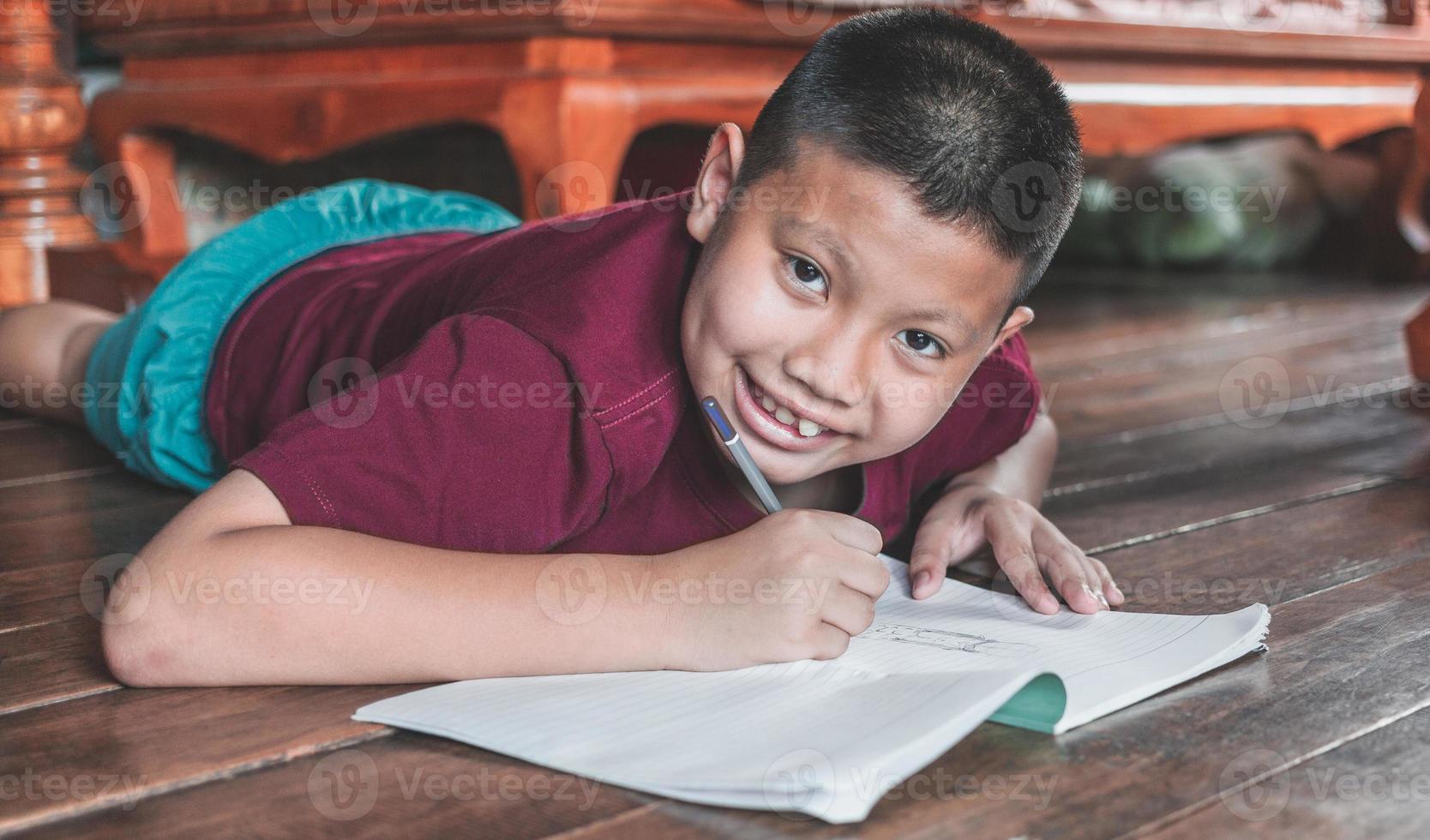 närbild av ett asiatisk pojke Sammanträde på de trä- golv skrivning en bok eller håller på med läxa med en leende roligt ansikte på Hem. foto