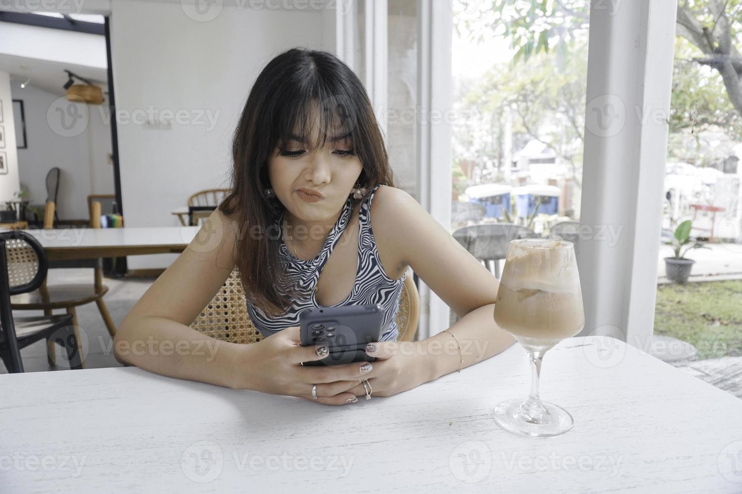 ledsen ansikte av en ung asiatisk kvinna innehav en cell telefon bär en tank topp Sammanträde nära en fönster. annons begrepp. foto
