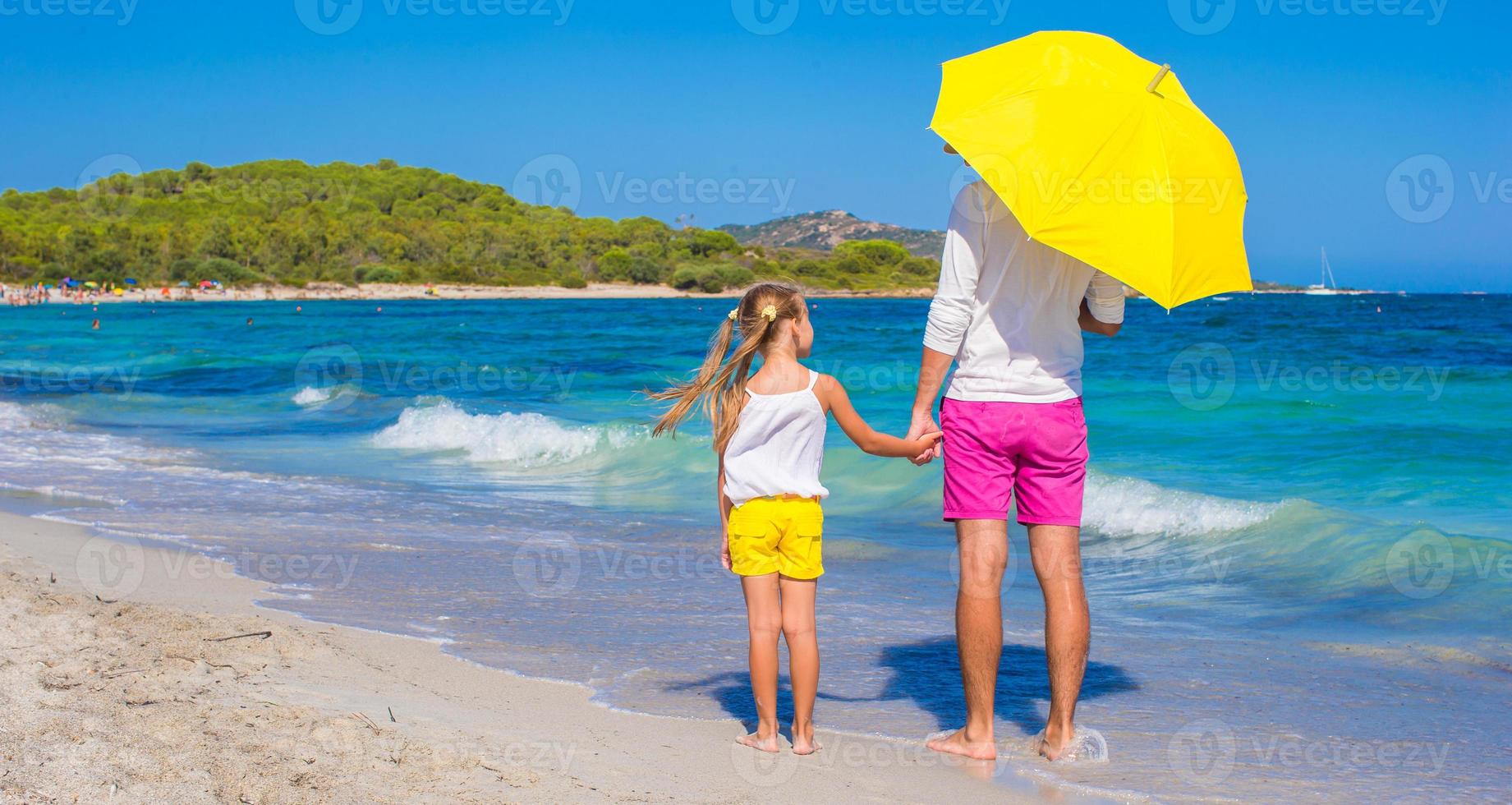 liten flicka och ung pappa på vit strand med gul paraply foto