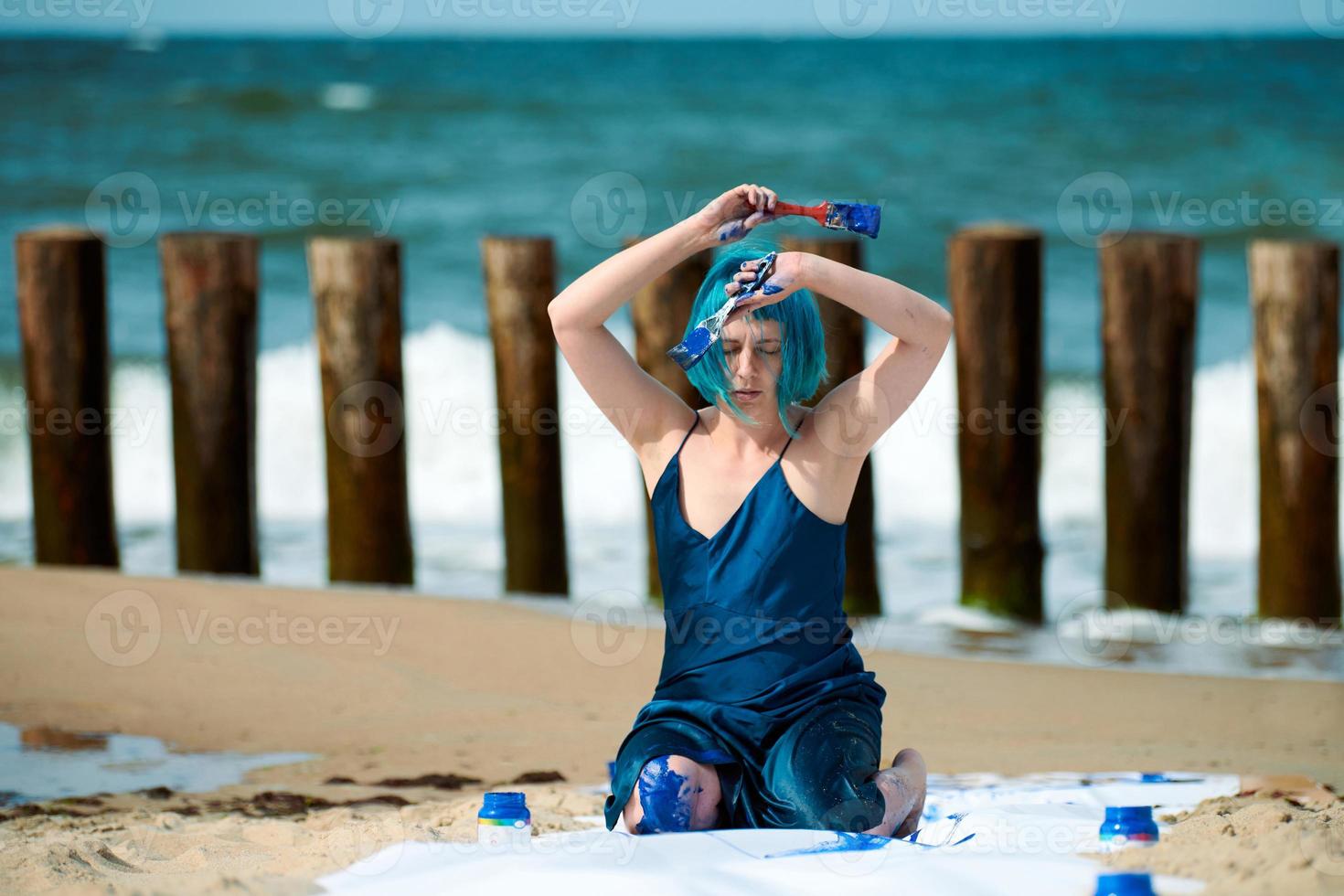 konstnärlig blåhårig kvinna prestanda konstnär insmord med blå gouache målarfärger Sammanträde på strand foto