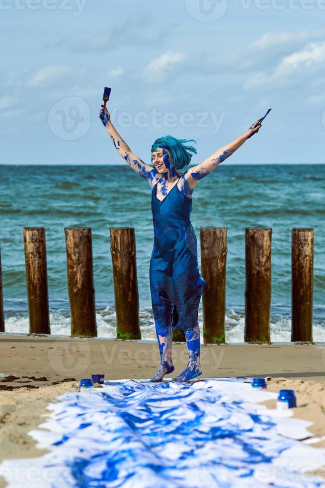 glad kvinna performance artist i blå klänning insmord med blå gouache färger dans på sandstrand foto