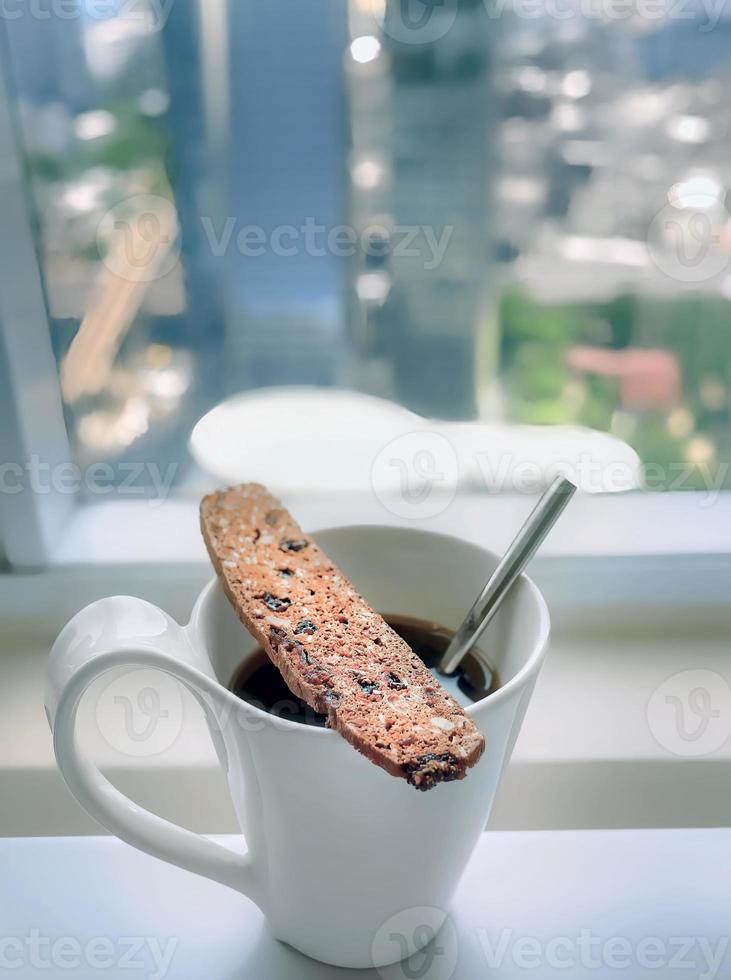 koreanska stil bild, en vit kopp av kaffe med biscotti, cantucci italiensk dubbel- bakad småkakor på topp, koppla av tid, kaffe ha sönder, med fönster suddig stad se i bakgrund foto