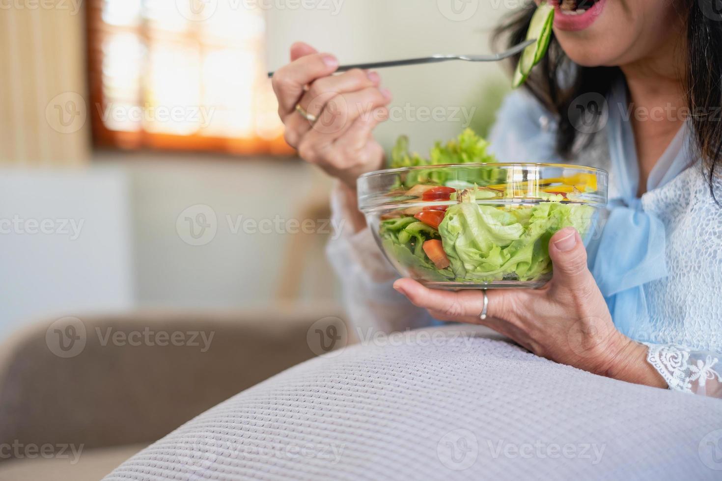 asiatisk senior kvinna Sammanträde äter vegetabiliska sallad och friska mat och äter lyckligt på de soffa i de hus för en friska kropp. friska mat begrepp foto