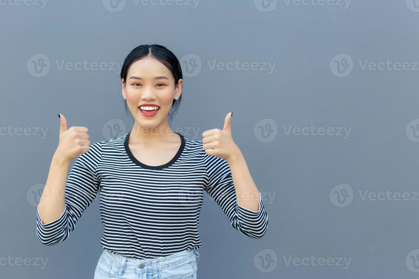 porträtt av söt asiatisk thai flicka studerande ser vänlig är stående leende lyckligt och självsäkert framgångsrik som visar dunk upp för kommunikation som Bra till närvarande något på en grå bakgrund. foto