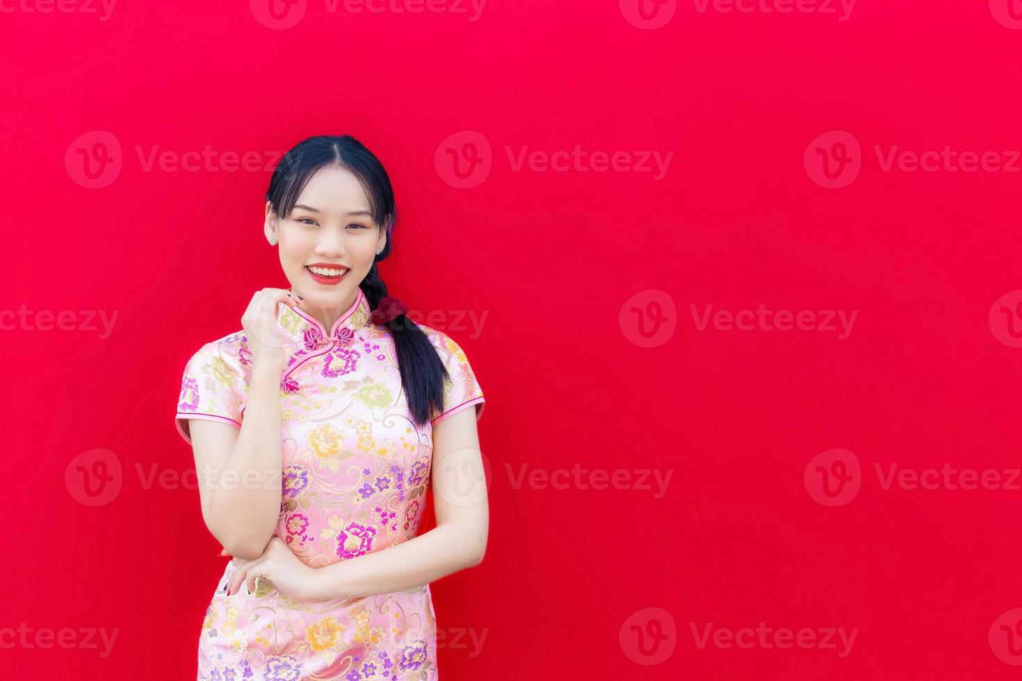 skön asiatisk kvinna bär rosa Cheongsam och ärm korsade medan utseende på till kamera och ler lyckligt med de röd bakgrund, fira kinesisk ny år tema. foto