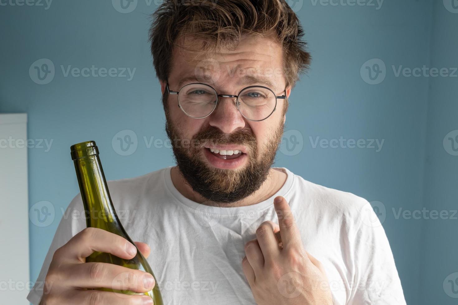 ovårdad man poäng hans finger på hans orakad. caucasian lider från en baksmälla, han innehar en vin flaska i händer och gör en grimas. foto