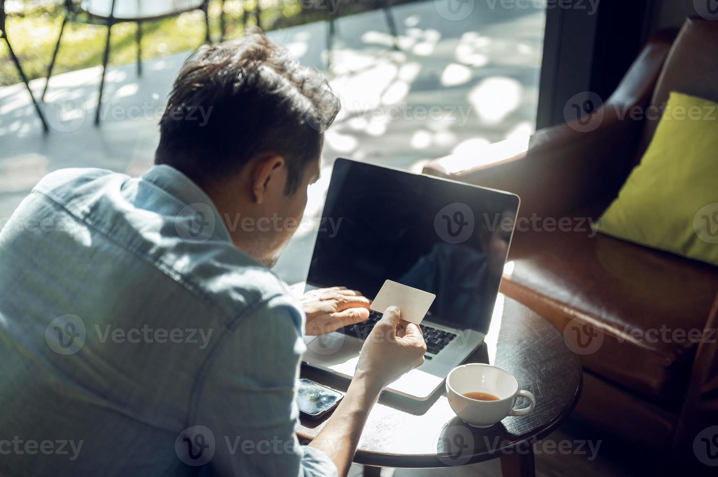 ung asiatisk man innehav kreditera kort och använder sig av bärbar dator uppkopplad betalning, uppkopplad handla begrepp med kreditera kort i Kafé foto