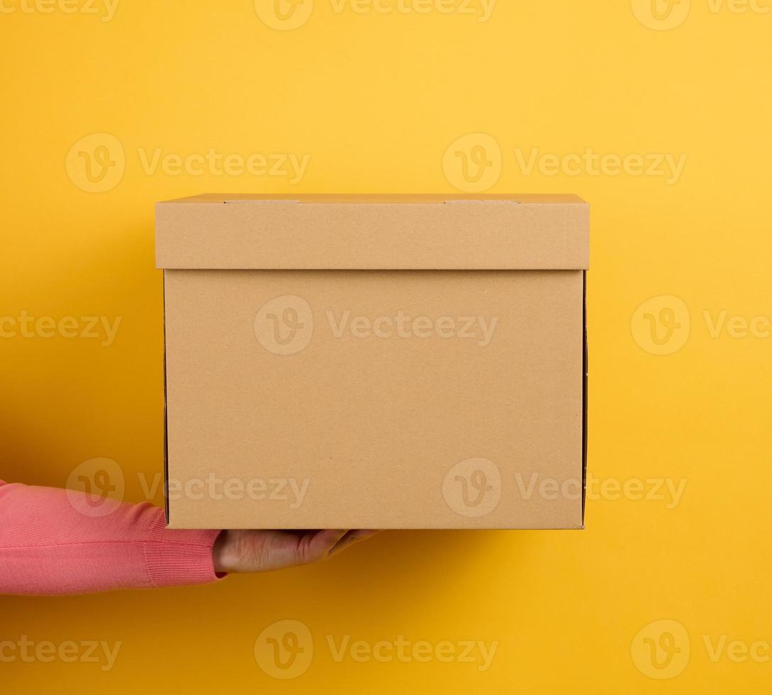kvinna hand innehav stängd brun kartong låda på gul bakgrund, leverans foto