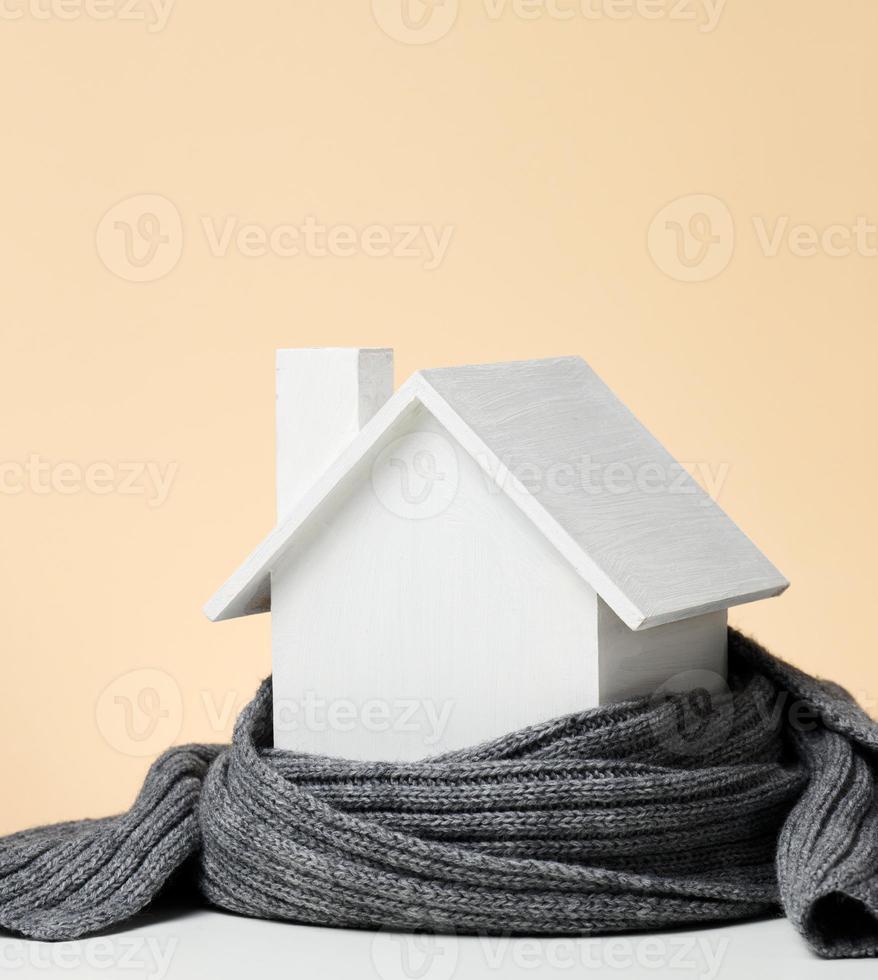 en vit trä- miniatyr- hus insvept i en grå stickat scarf. byggnad isolering begrepp, lån för reparationer foto