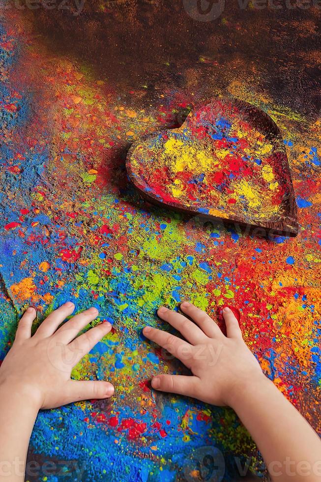 barns händer lögn på en tabell på som holly's målarfärger är spridd och en trä- tallrik i de form av en hjärta, i som ljus färger, kopia Plats. indisk festival av färger holi foto