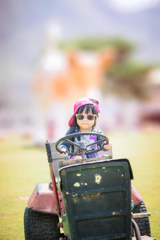 söt asiatisk liten flicka bär solglasögon som spelar genom att köra en gammal traktor på gården foto
