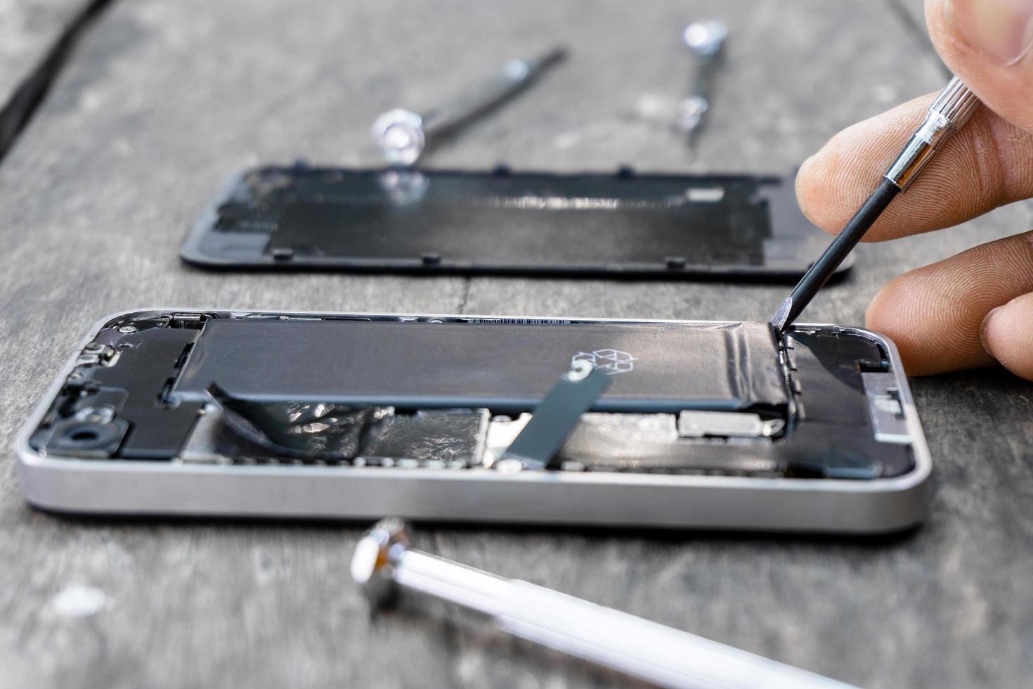 teknikern håller en skruvmejsel en mobiltelefon reparation närbild inuti mobiltelefon med ett fixeringsbatteri från ett trasigt servicecenter på träbord. smartphone reparationer underhåll koncept. foto