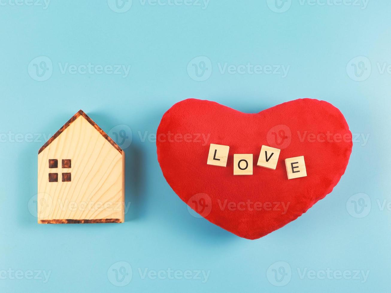 platt layout av trä- modell hus och röd hjärta form kudde med trä- brev l o v e isolerat på blå bakgrund med kopia Plats, valentines eller Hem av kärlek begrepp. foto