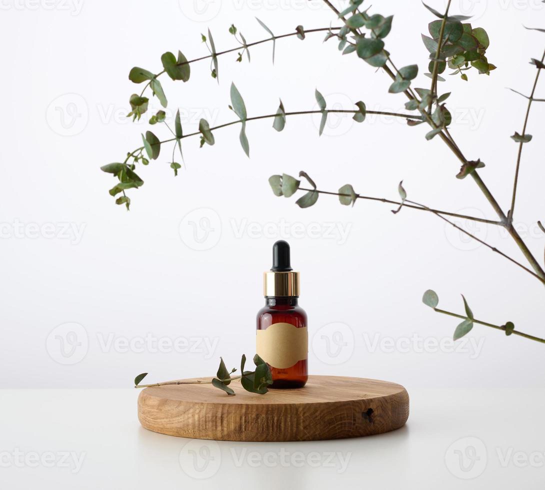 en glas brun flaska med en pipett på en trä- podium och en gren av eukalyptus, vit bakgrund. reklam och befordran av kosmetisk Produkter foto