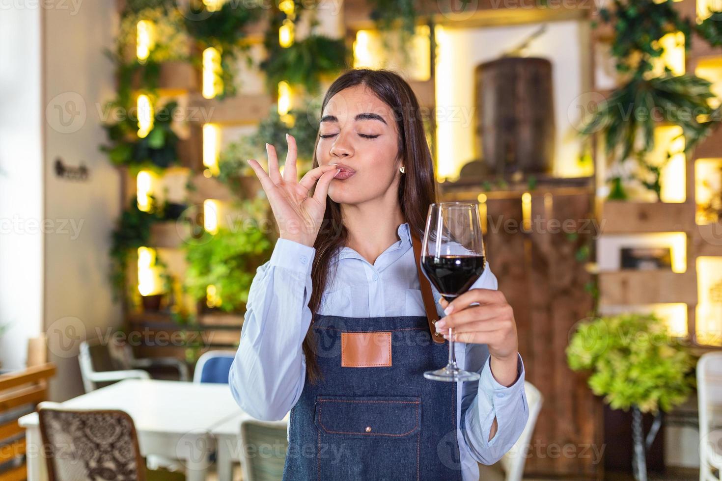 glad elegant servitris innehav glas av röd vin som visar utsökt tecken , stående i restaurang. sommelier provsmakning vin i restaurang. foto