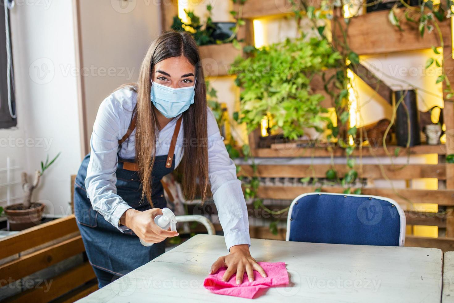 porträtt av attraktiv kvinna servitris bär ansikte mask och rengöring de tabell med alkohol och våt våtservetter innan Välkommen kund. ny vanligt med coronavirus utbrott hygien restaurang begrepp. foto