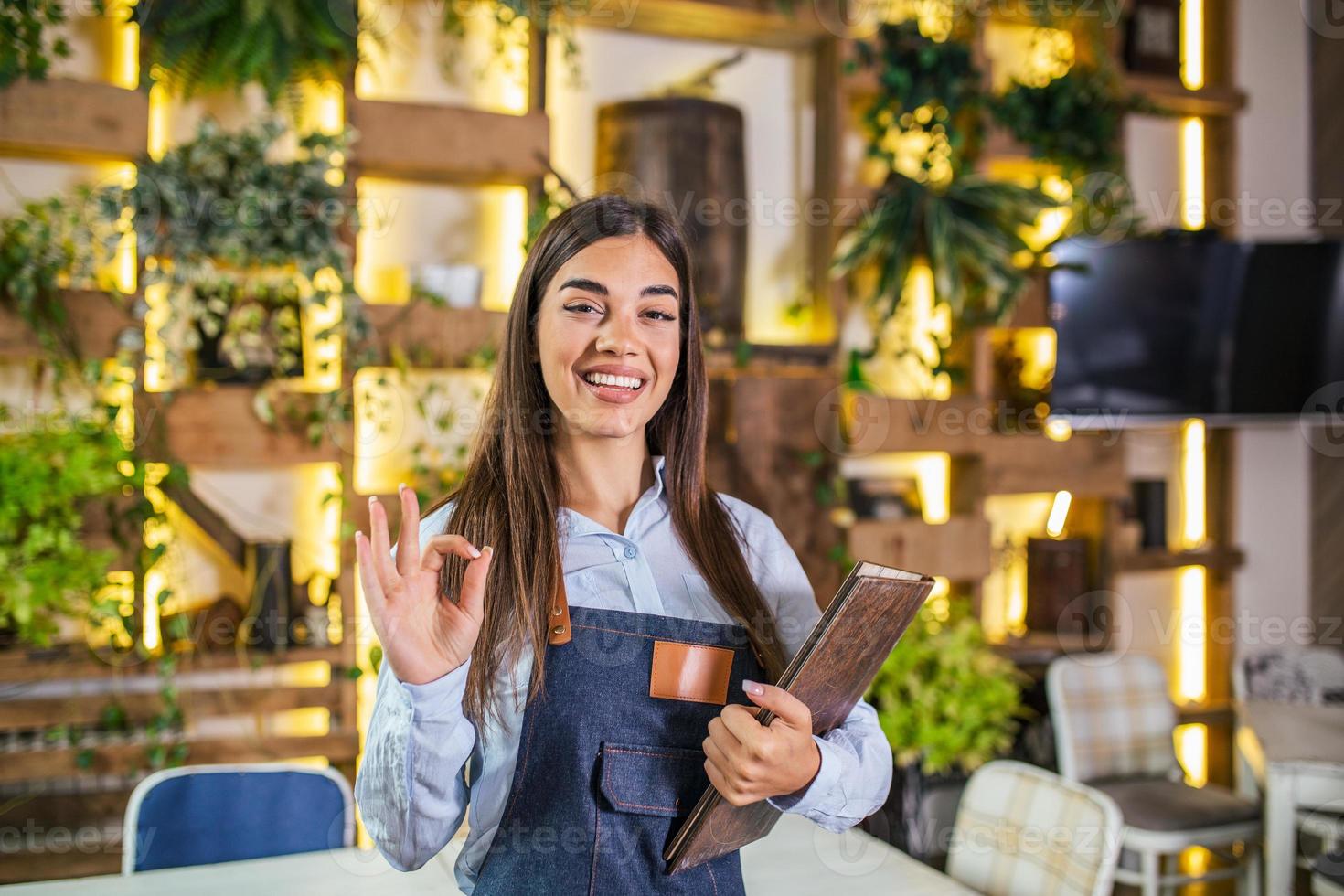 skön kvinna servitris leende i de restaurang innehav en meny och som visar ok tecken. foto