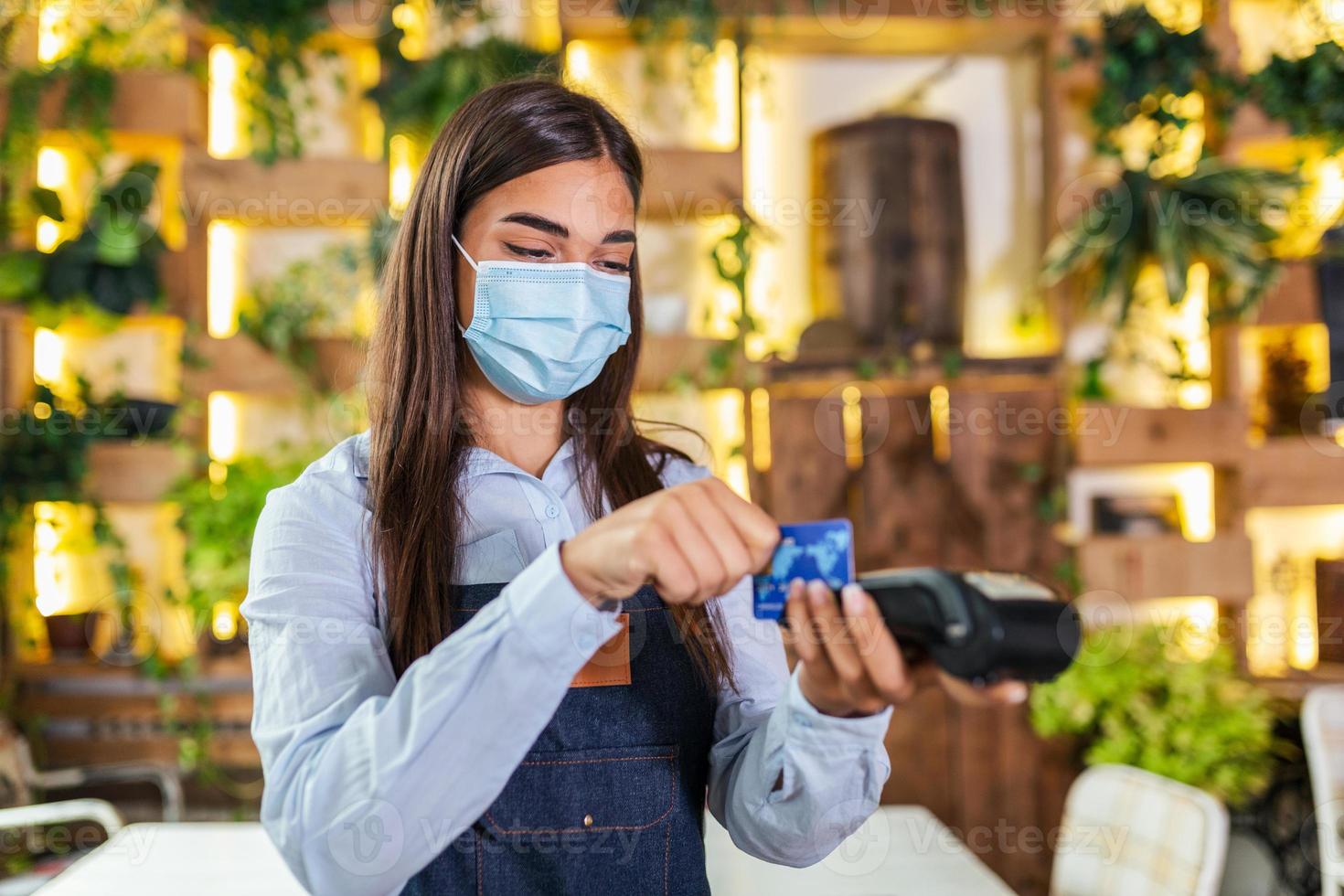 servitris med skyddande ansikte mask framställning en kreditera kort betalning med kort läsare maskin. kreditera kort slägga genom terminal i restaurang. servitris hand svep debitera kort till betala de räkningen foto