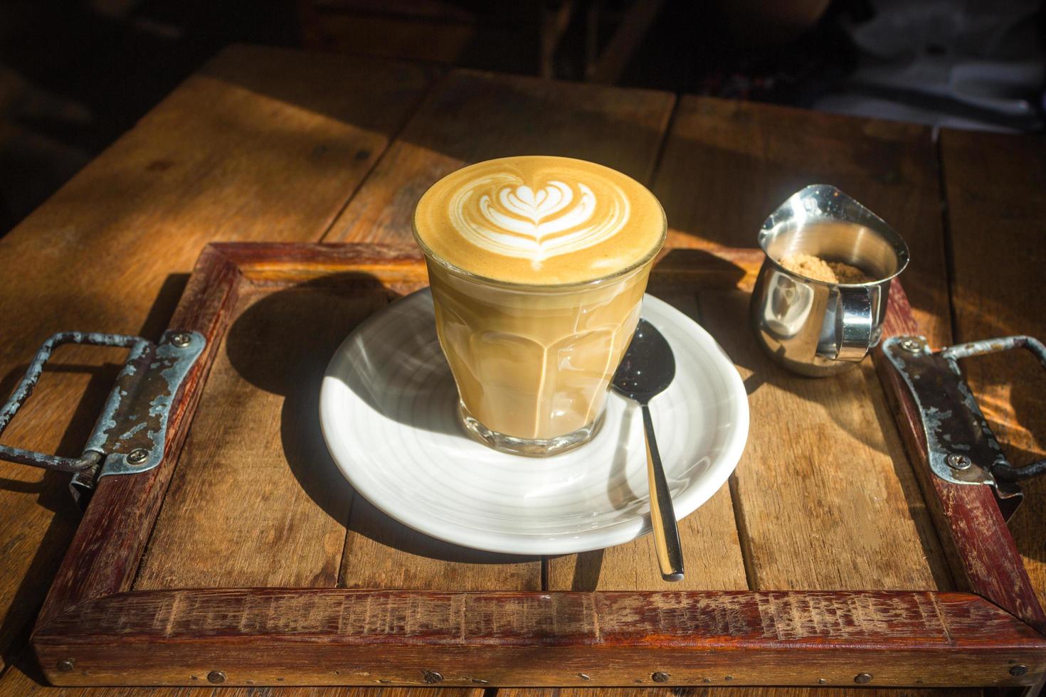 kaffe med lattekonst på vit platta och träbricka i solljus foto