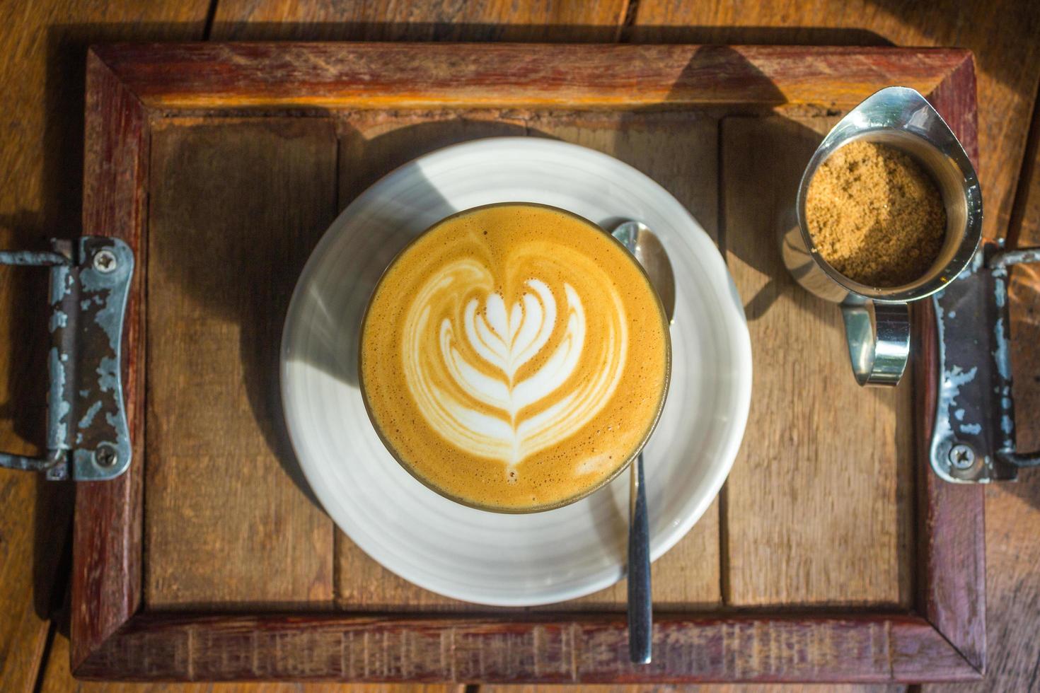 kaffe med lattekonst på vit platta och träbricka i solljus foto