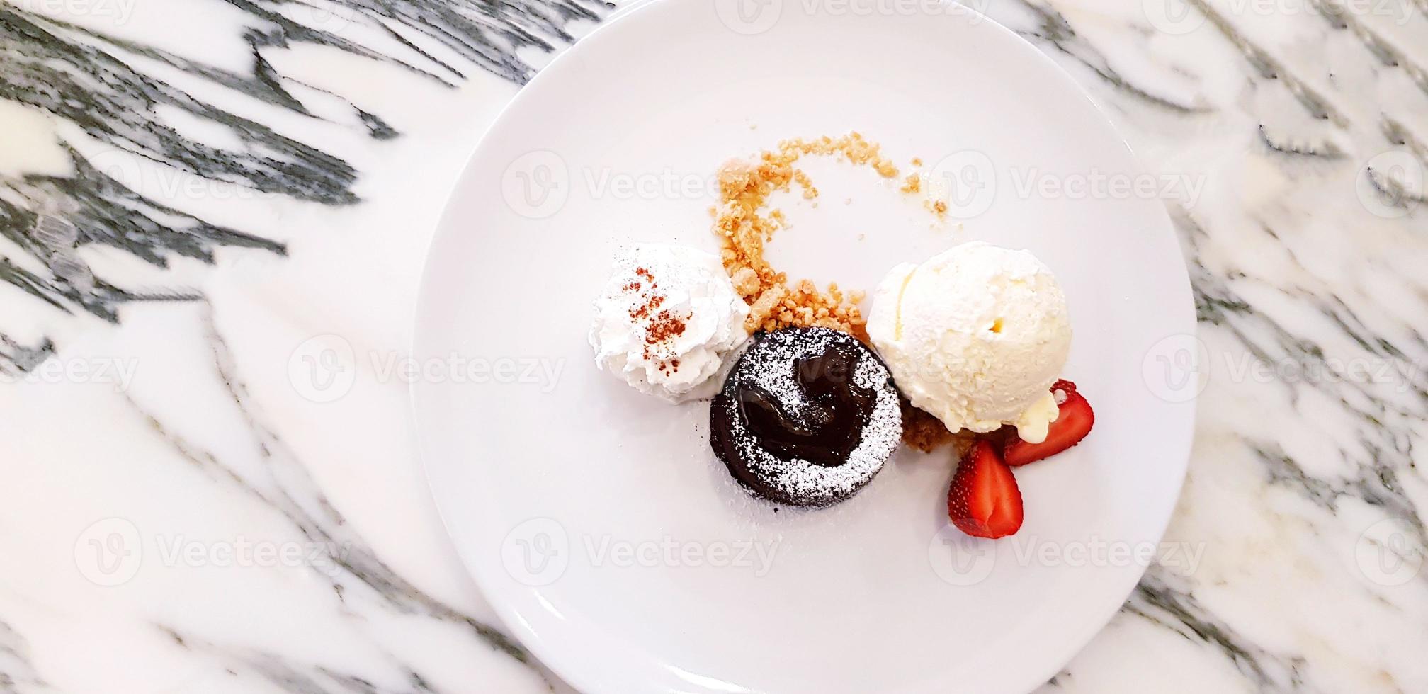 platt lägga eller topp se av choklad lava kaka med vanilj is grädde, vispad grädde och skivad av jordgubb i tallrik på vit marmor tabell. efterrätt och ljuv mat begrepp foto