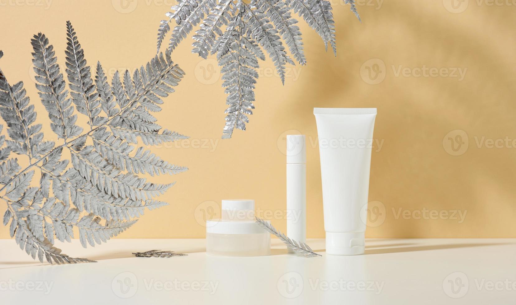 vit rör för kosmetika, en burk av grädde och silverren på en vit tabell. kosmetisk på en beige skugga bakgrund. grädde flaska, lotion, rengöringsmedel, schampo för hudvård foto
