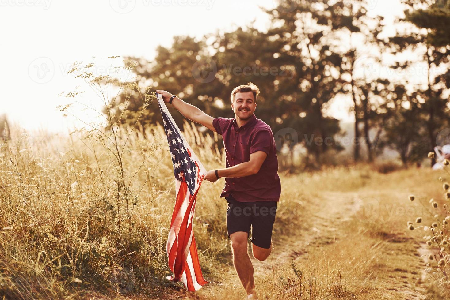 vuxen man kör med amerikan flagga i händer utomhus i de fält. känner frihet på solig dagtid foto