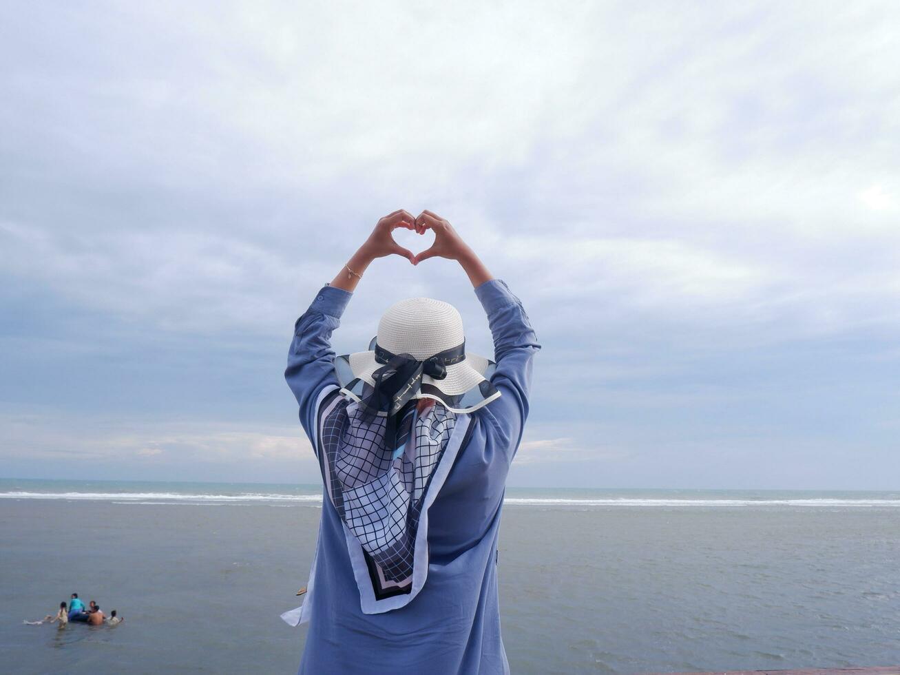 tillbaka av de kvinna i de hatt på de tropisk strand vem är ser upp på de himmel och de hav medan henne händer form kärlek foto